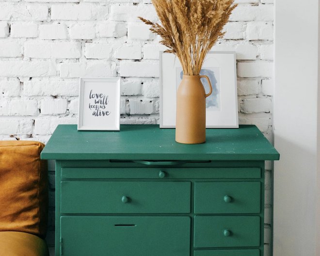 A green chest of drawers with two squares, one with a cursive text and the other one is behind the vase on top of the chest of drawers.